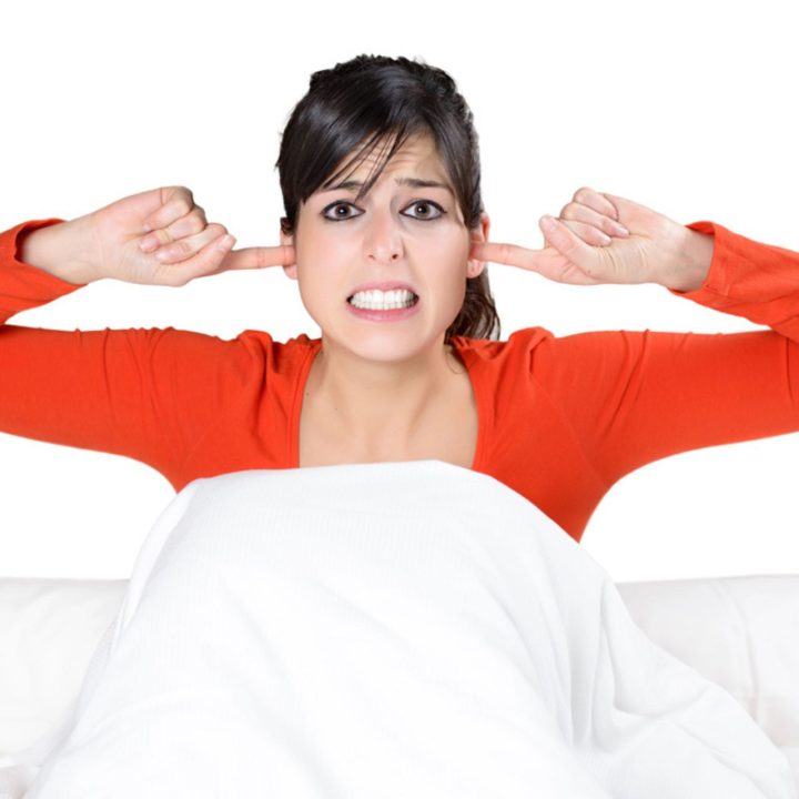 Femme stressée dans son lit, les doigts dans les oreilles à cause du bruit extérieur – illustration des nuisances sonores et de l'importance de l'isolation acoustique des fenêtres.
